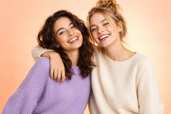 The same two cheerful friends pose against a softly lit peach and cream studio background.