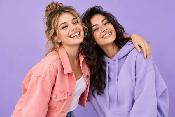 Two cheerful friends in colorful casual clothes pose together against a lavender studio background.